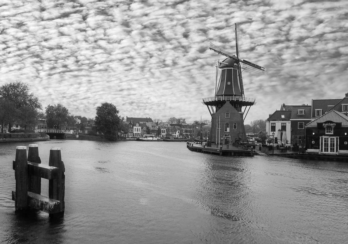 De Adriaan — Haarlems bekendste molen aan het Spaarne, de wieken afgetekend tegen een schapenwolkenlucht, met trapgevels en oude meerpalen langs de stille rivier — in zwart-wit.