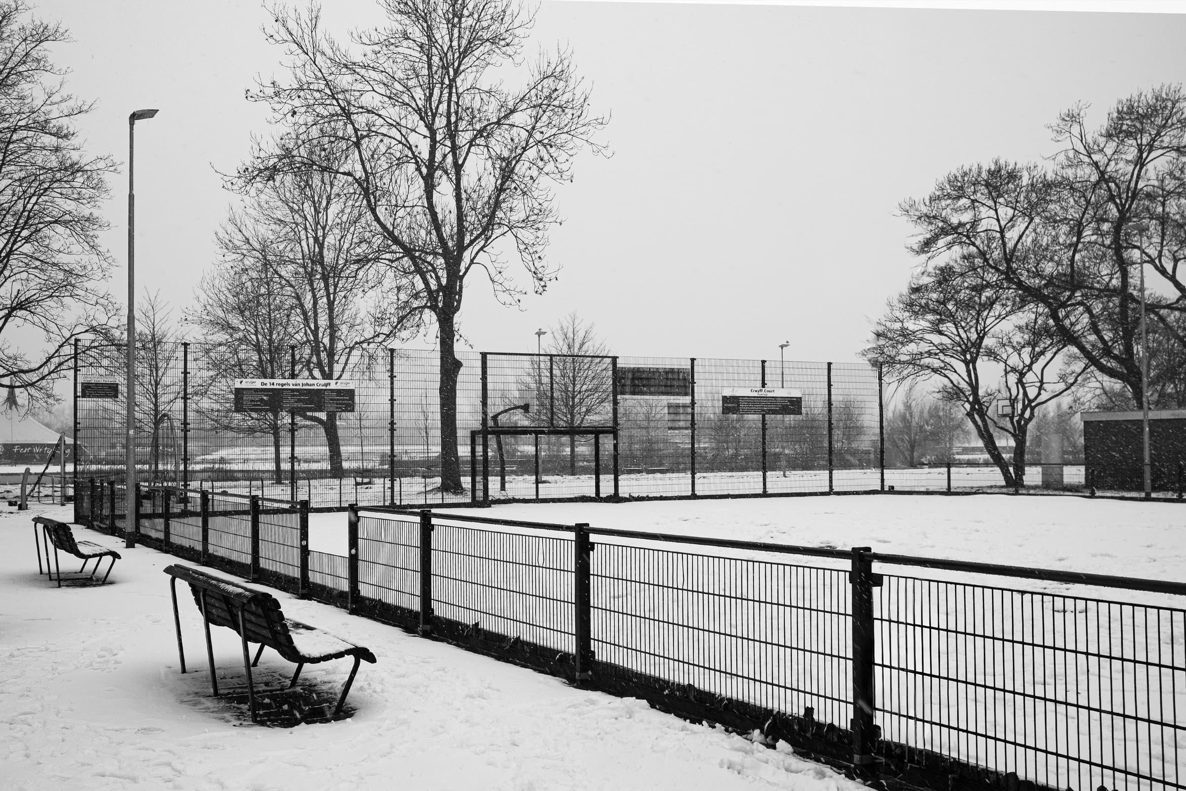 Off Season — Een leeg, besneeuwd Cruyff Court ligt er stil bij achter het hekwerk, met kale bomen en verlaten bankjes onder een egaal grijze lucht — een speelveld tot stilstand gebracht door de winter, in zwart-wit.