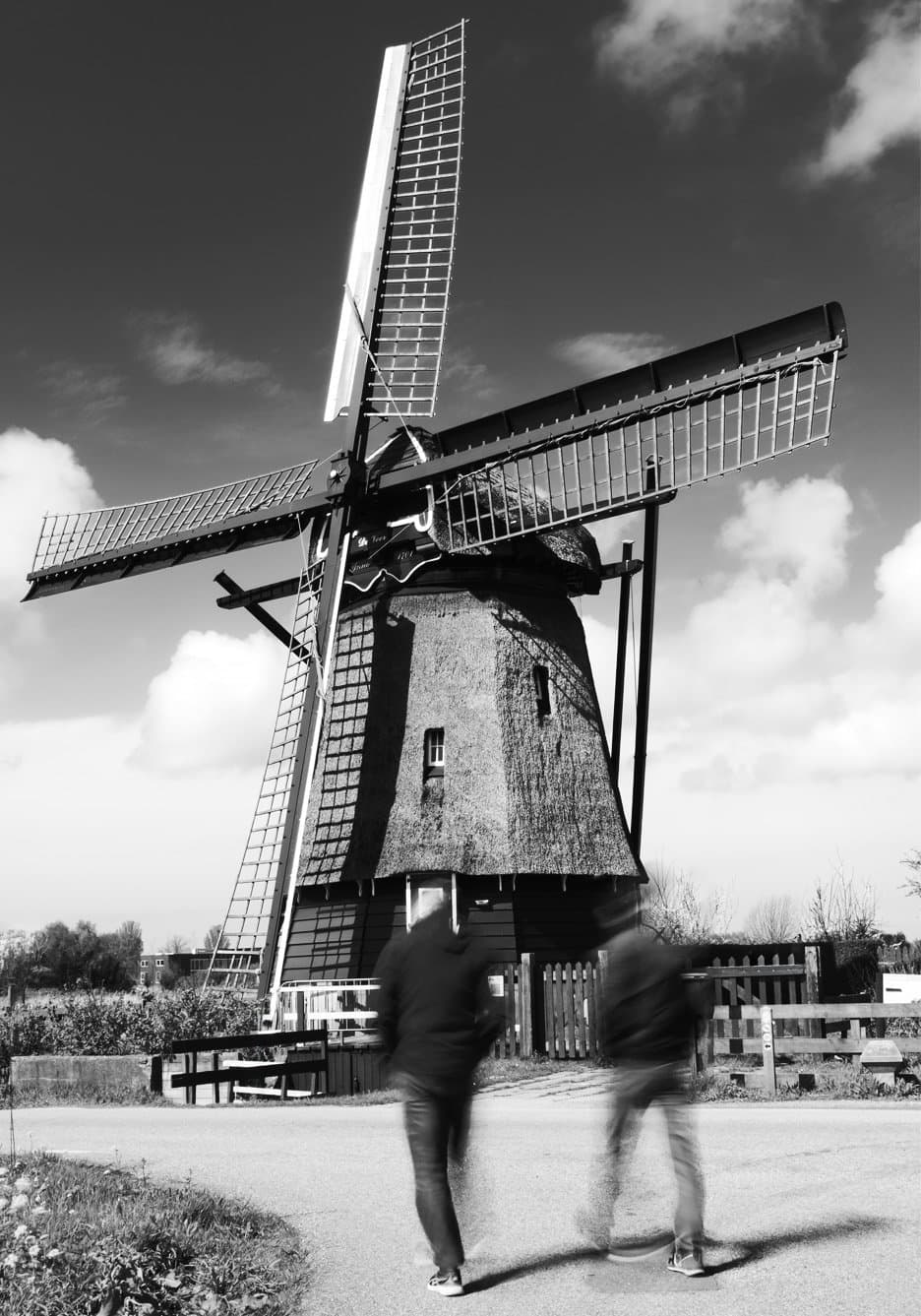 Passing By — Een rietgedekte molen torent tegen een heldere, wolkenrijke lucht met wijd gespreide wieken, terwijl een bewogen figuur langs de bankjes op het pad beneden loopt — stilte en beweging samen gevangen in zwart-wit.