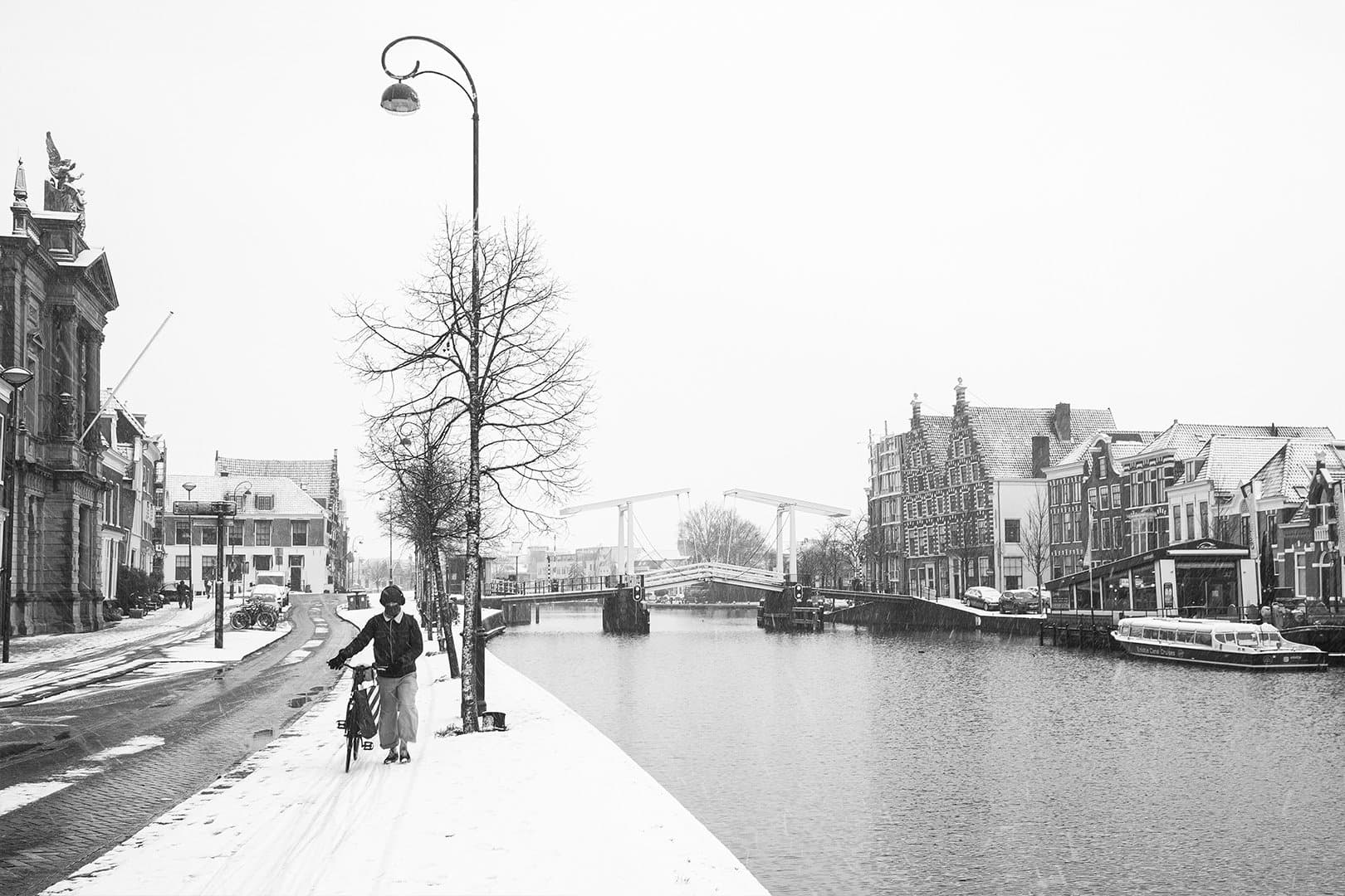 First Snow — Een eenzame figuur loopt met een fiets over een besneeuwde kade langs de gracht in Haarlem, terwijl de witte ophaalbrug en trapgevels vervagen in een bleke winterlucht — stil en roerloos in zwart-wit.